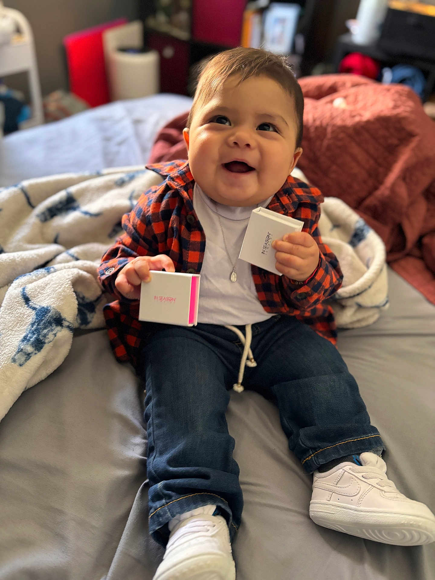 Paxton is registered to the contest to win money with this photo: baby, child, smiling, jeans, shirt, white_shoes, blanket, bed, indoors, person, happy, clothing, plaid, sitting, cute, holding, toy, casual, footwear, portrait