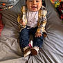 Paxton is registered to the contest to win money with this photo: toddler, child, smiling, sitting, plaid_jacket, jeans, white_shirt, brown_shoes, bed, blanket, toy, remote_control, room, happy, indoor, clothing, face, person, cute, playful