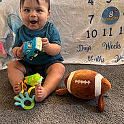 Paxton is registered to the contest to win money with this photo: baby, child, toy, plush_toy, football, carpet, milestone_blanket, smiling, sitting, blue_shirt, shorts, indoor, floor, cute, face, hand, foot, playing, happy, infant