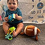 baby, child, toy, plush_toy, football, carpet, milestone_blanket, smiling, sitting, blue_shirt, shorts, indoor, floor, cute, face, hand, foot, playing, happy, infant
