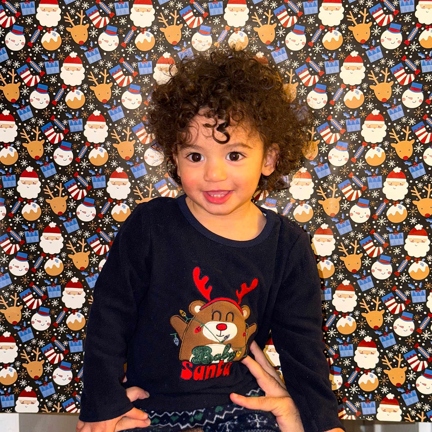 Youssef Ghali participe au concours pour gagner de l'argent avec cette photo : adult_hands, celebration, child, christmas, clothing, curly_hair, cute, face, festive, happy, holiday, indoors, patterned_background, person, portrait, reindeer_sweater, santa, shoulders, smiling, toddler