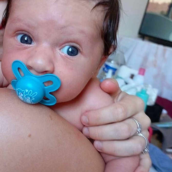 Isma participe au concours pour gagner de l'argent avec cette photo : adult, baby, baby_holding, blue_pacifier, close_up, comfort, expression, eyes, face, hand, human, indoor, infant, pacifier, person, portrait, resting, room, shoulder, skin