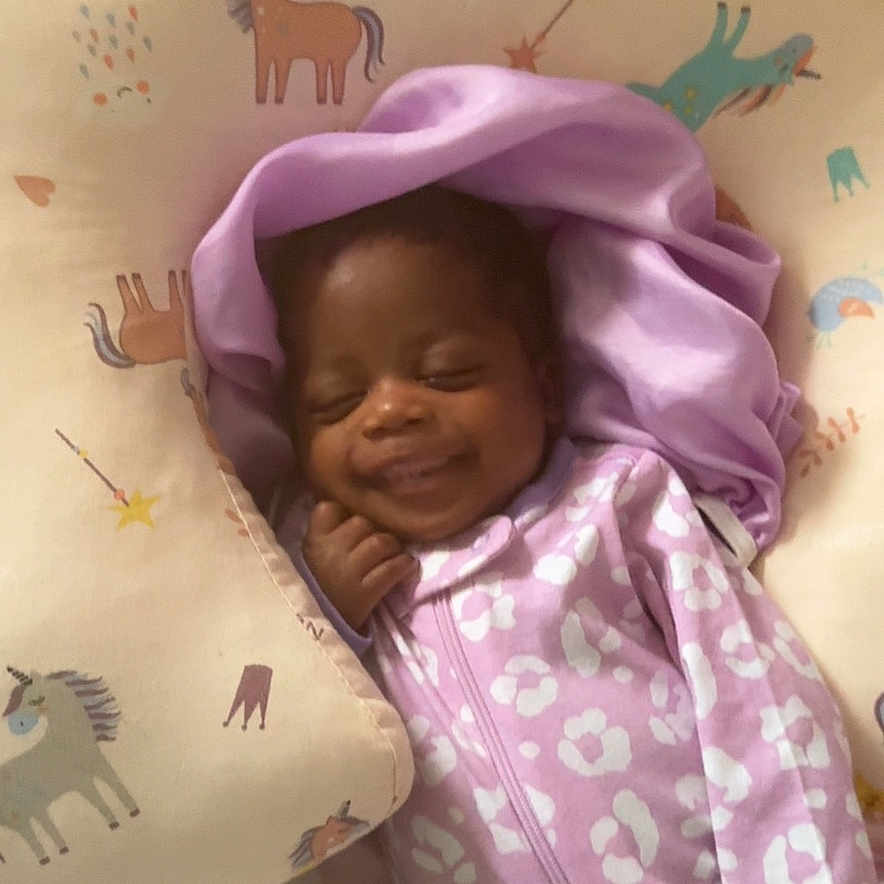 Nadirah is registered to the contest to win money with this photo: baby, bed, blanket, child, comfort, cozy, cushion, cute, face, happy, indoors, infant, onesie, person, purple_clothing, resting, sleeping, smiling, soft_fabric, unicorn_pattern