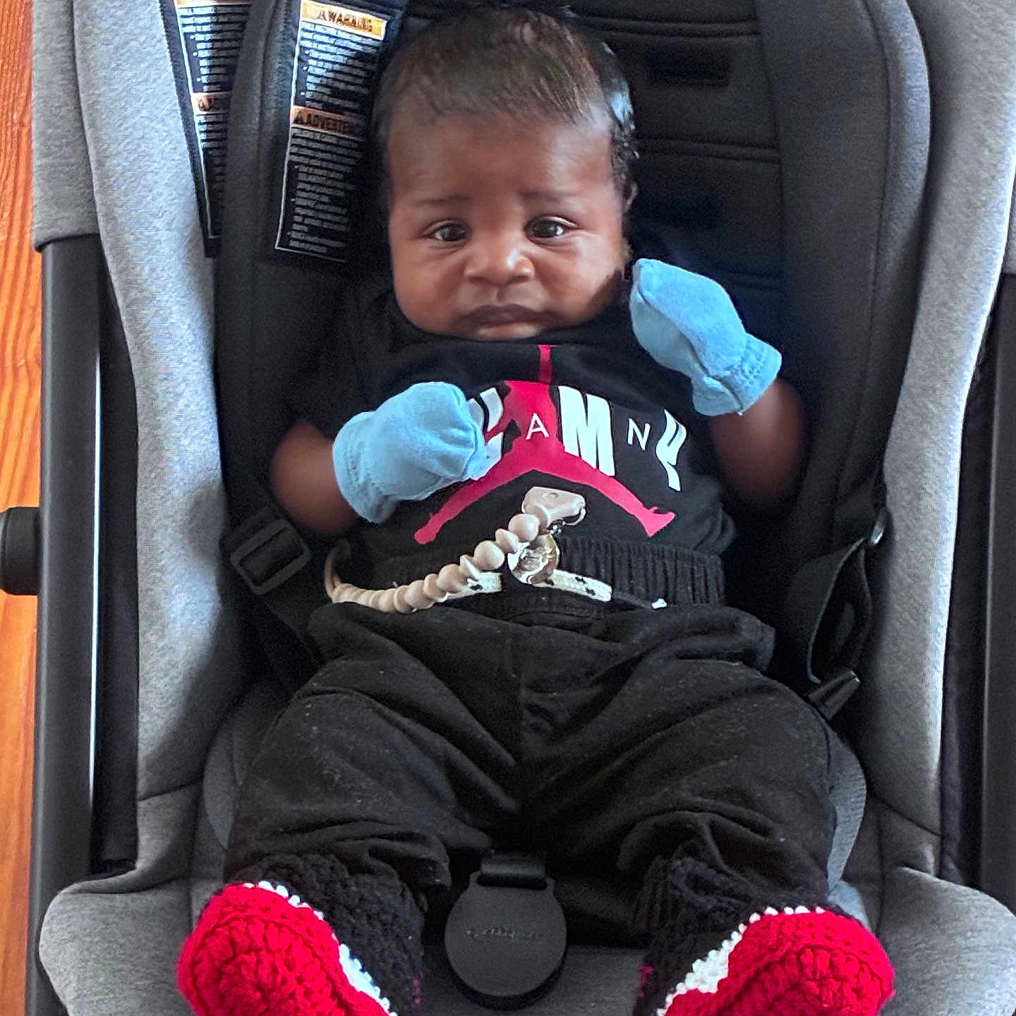 Travis is registered to the contest to win money with this photo: baby, infant, car_seat, clothing, mittens, black_clothes, red_booties, knitted_booties, baby_jogger, seat_belt, person, portrait, indoor, child, cute, young, face, head, sitting, expression