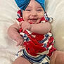 baby, smiling, blue_bow, red_clothing, white_blanket, happy, infant, child, cute, portrait, close_up, chubby_arms, candid, indoors, soft_texture, colorful_outfit, cheerful, person, headwear, playful