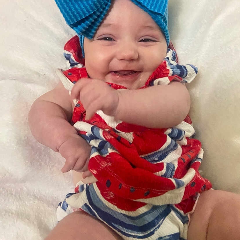 Blakelee is registered to the contest to win money with this photo: baby, blue_bow, candid, cheerful, child, chubby_arms, close_up, colorful_outfit, cute, happy, headwear, indoors, infant, person, playful, portrait, red_clothing, smiling, soft_texture, white_blanket