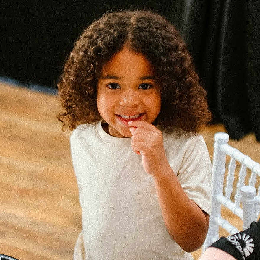 Jakobi joined the competition — help win amazing prizes! child, curly_hair, smiling, white_shirt, indoor, chair, table, person, happy, young, portrait, wooden_floor, casual_clothing, bright, cute, playful, people, friends, light_skin, seated