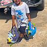 child, boy, easter_basket, plush, braided_hair, tshirt, shorts, sneakers, sunlight, outdoor, driveway, vehicle, license_plate, concrete, smiling, holding, toy, holiday, spring, daytime