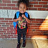 child, smiling, brick_wall, backpack, black_clothing, toddler, standing, outdoor, concrete_floor, dinosaur_tshirt, sneakers, happy, curly_hair, sunlight, shadow, person, casual, fashion, portrait, daylight