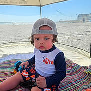 Brayden is registered to the contest to win money with this photo: toddler, child, beach, tent, hat, cap, swimsuit, fish_pattern, toy, blanket, sunglasses, sand, ocean, outdoor, summer, sun_protection, colorful, striped, serious_expression, mesh