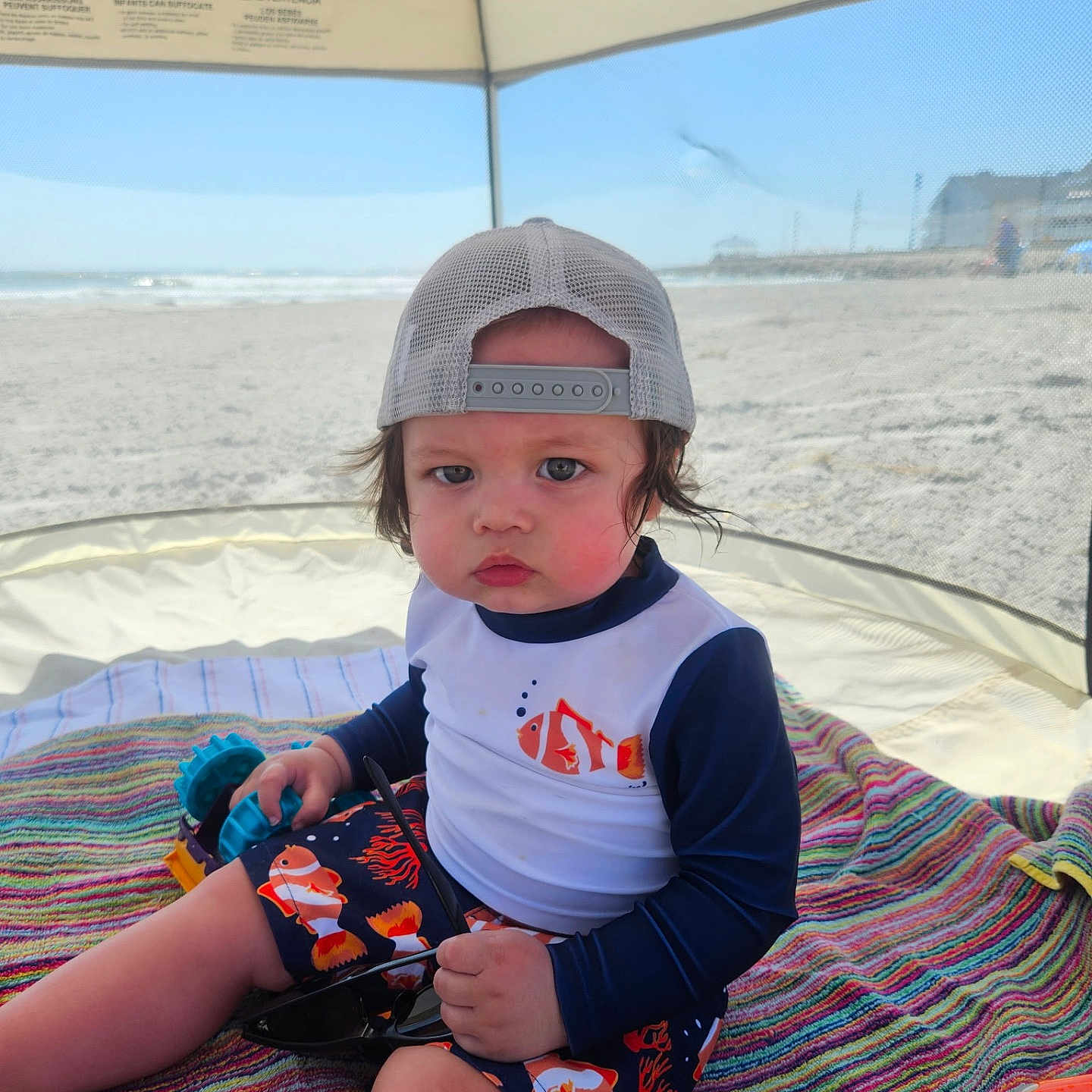 Brayden is registered to the contest to win money with this photo: toddler, child, beach, tent, hat, cap, swimsuit, fish_pattern, toy, blanket, sunglasses, sand, ocean, outdoor, summer, sun_protection, colorful, striped, serious_expression, mesh