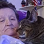 elderly_person, tabby_cat, indoor, sitting, close_up, face, pet, cat_print, shelf, american_flag, portrait, home_decor, relaxing, furniture, comfort, feline, casual_clothing, photo_frame, lamp, snuggling