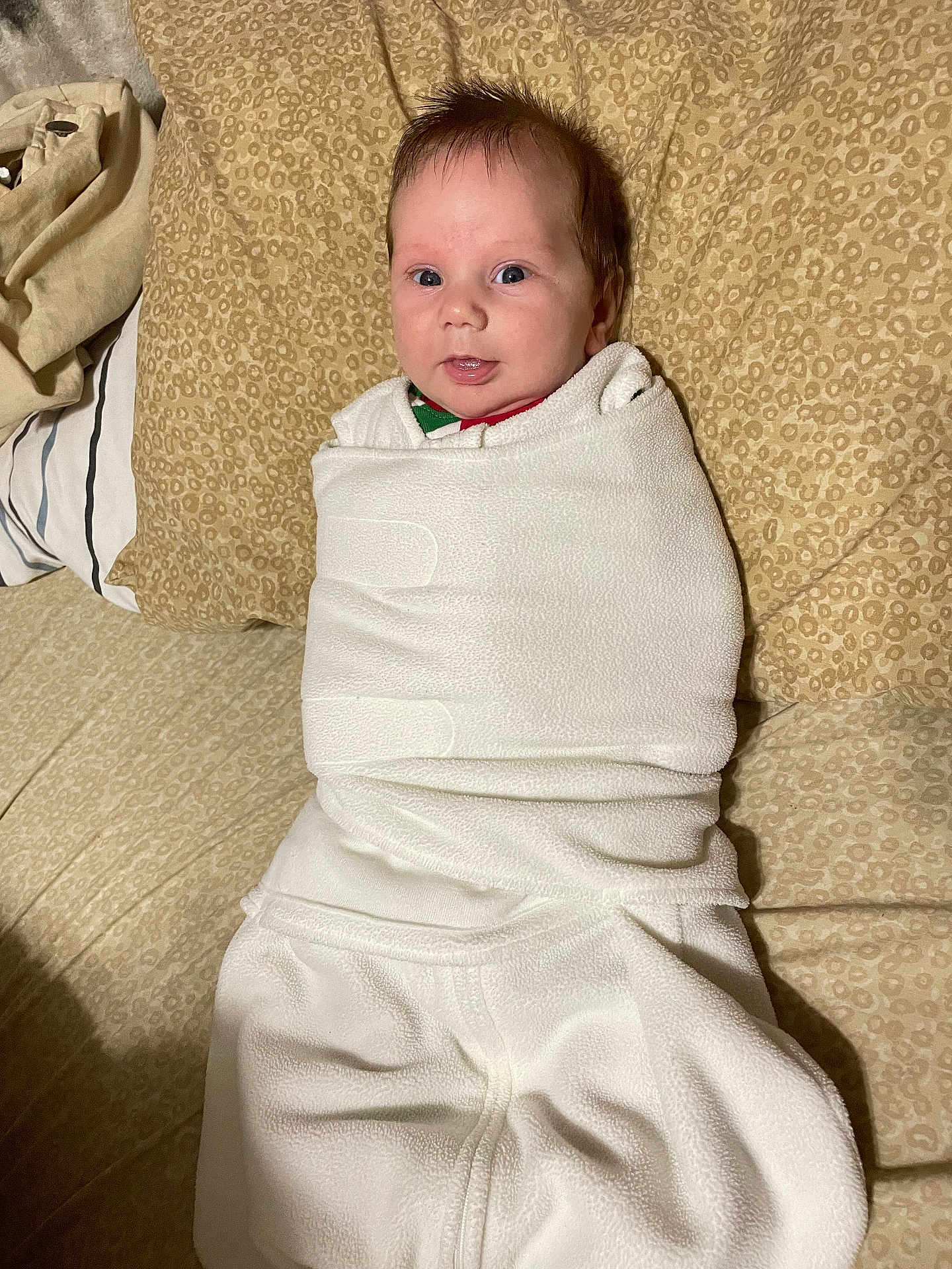 Benjamin is registered to the contest to win money with this photo: baby, infant, blanket, white_clothing, bed, pillow, patterned_bedspread, face, eyes, smile, wrapped, cozy, indoors, child, newborn, cute, soft_texture, lying_down, portrait, home