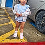 child, toddler, parking_lot, car, open_car_door, blue_door, sandals, socks, dress, jacket, pavement, wheel, parking_lines, red_paint, concrete, building, balcony, portrait, outdoor, cute