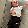 baby, infant, child, white_onesie, black_pants, nike_logo, socks, couch, cushion, fabric, indoor, person, cute, portrait, small_child, looking_up, relaxed, lying_down, soft_lighting, casual_clothing