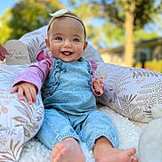 Jordyn is registered to the contest to win money with this photo: baby, infant, smiling, blue_eyes, headband, denim_overalls, pink_shirt, barefoot, cushion, blanket, plush_toy, outdoor, sunlight, happy, sitting, portrait, child, soft_texture, nature, blurred_background