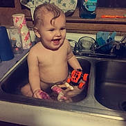 Ava is registered to the contest to win money with this photo: baby, child, sink, bath, water, toy, kitchen, smiling, happy, towel, dish_soap, window, cabinet, curly_hair, cute, indoor, playful, cleaning, plastic_toy, splashing