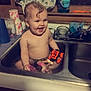 baby, child, sink, bath, water, toy, kitchen, smiling, happy, towel, dish_soap, window, cabinet, curly_hair, cute, indoor, playful, cleaning, plastic_toy, splashing