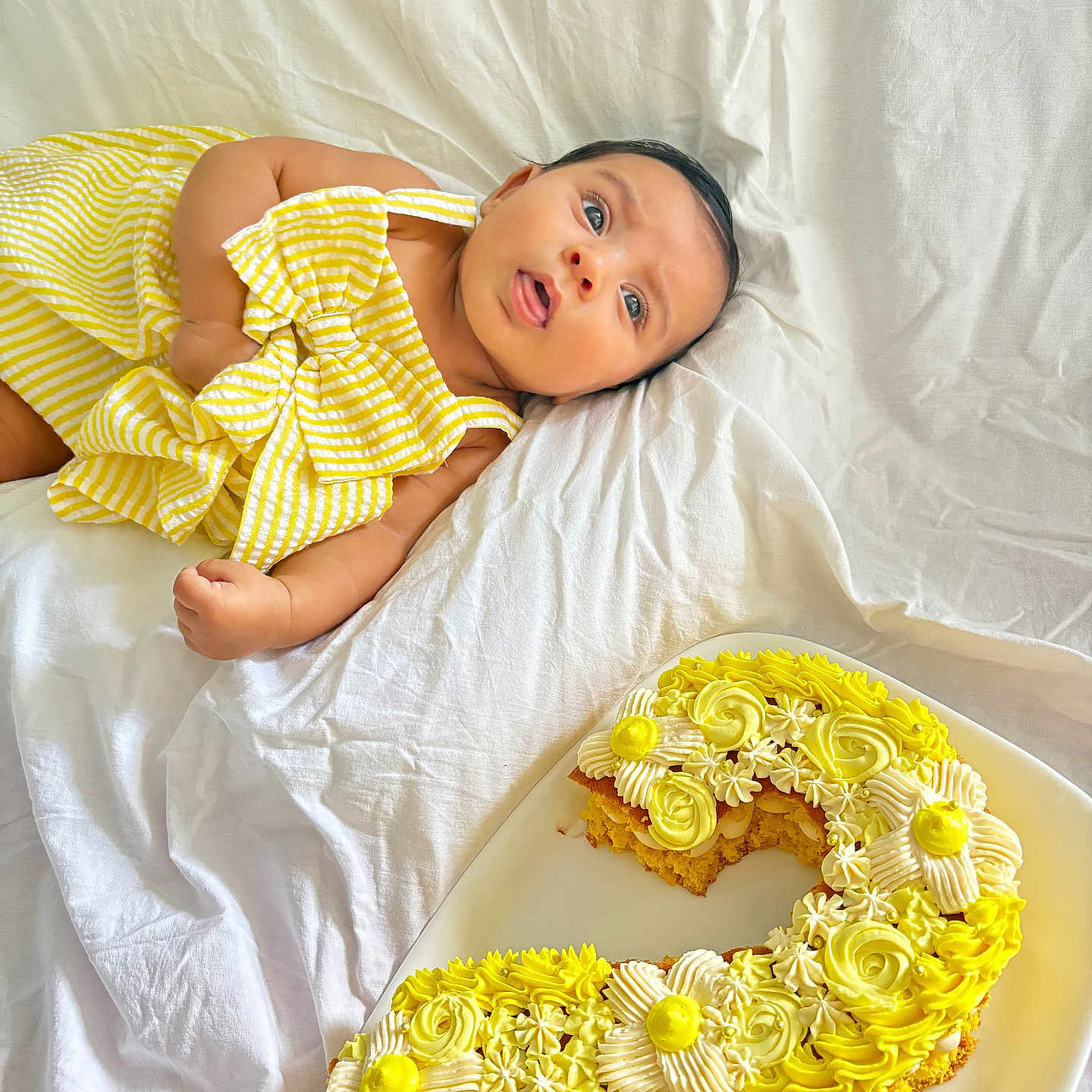 Leylani a rejoint le concours — aidez-le/la à gagner de superbes lots ! baby, birthday_cake, cake, celebration, child, cute, decorated_cake, food, frosting, icing, infant, lying_down, number_two_cake, person, plate, portrait, striped_clothing, sweet, white_sheet, yellow_dress