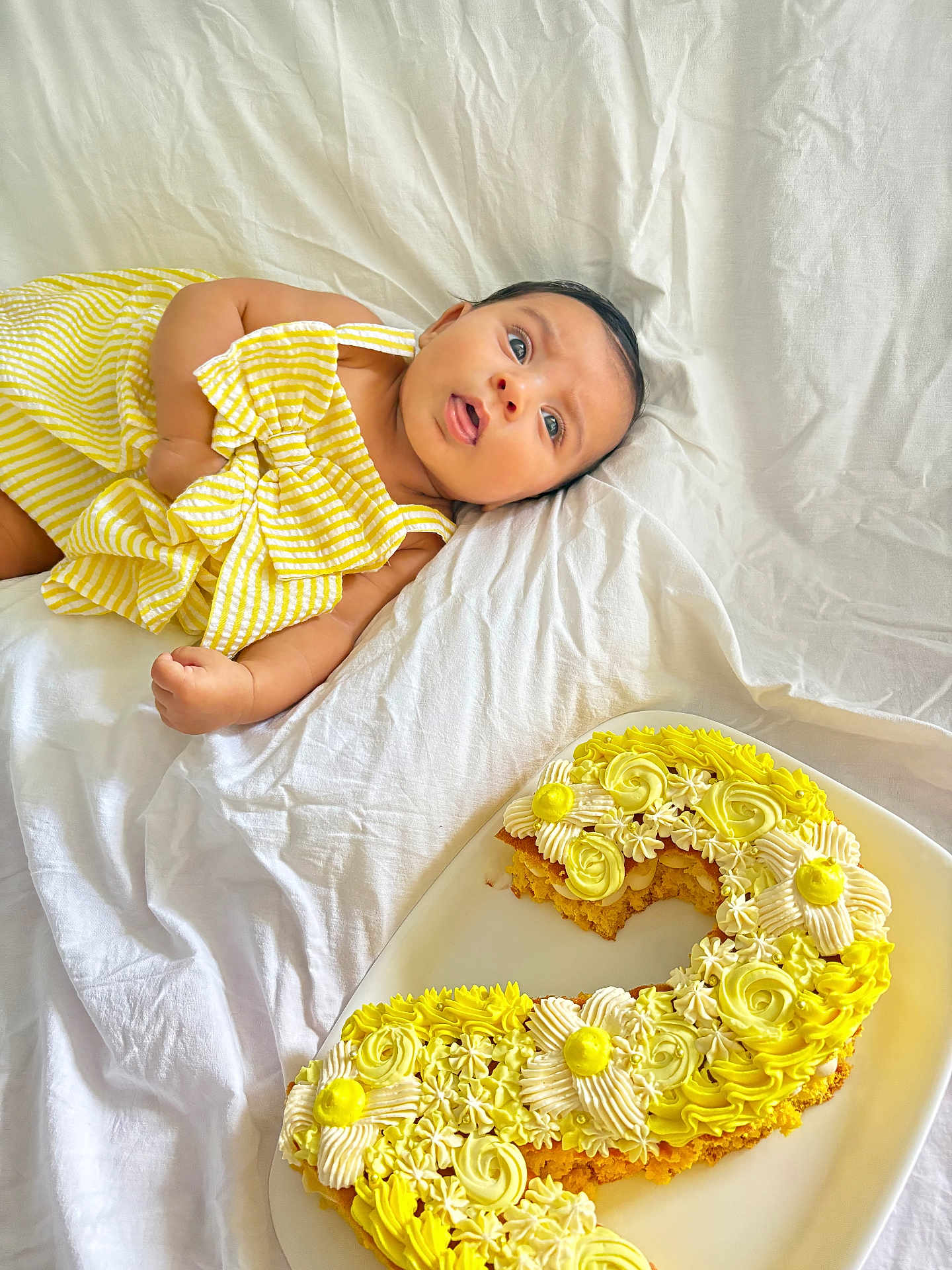 Leylani a rejoint le concours — aidez-le/la à gagner de superbes lots ! baby, birthday_cake, cake, celebration, child, cute, decorated_cake, food, frosting, icing, infant, lying_down, number_two_cake, person, plate, portrait, striped_clothing, sweet, white_sheet, yellow_dress