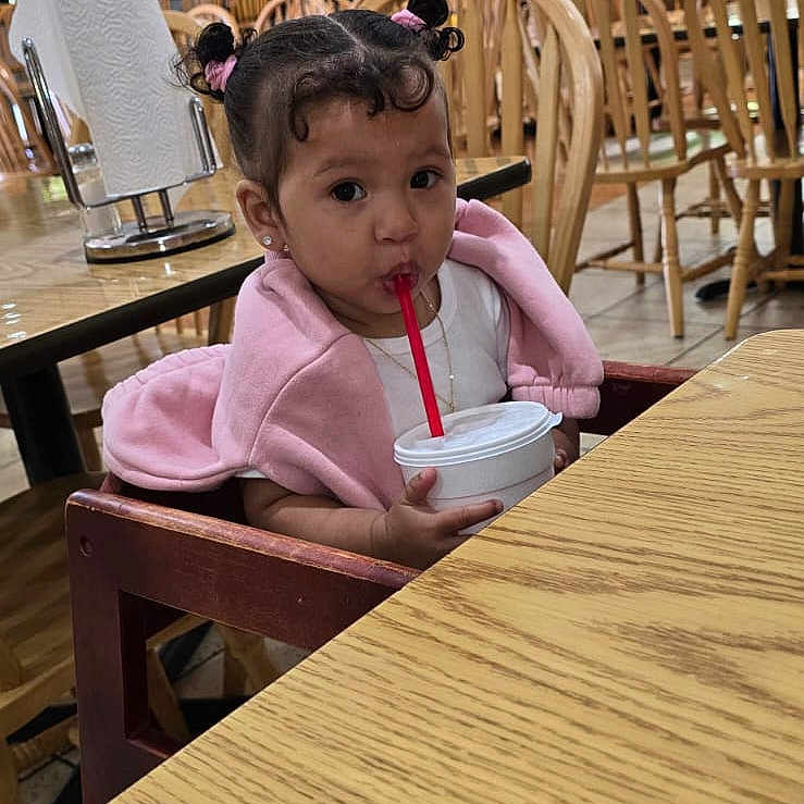 Rachell Mendoza is registered to the contest to win money with this photo: casual_clothing, child, cup, curly_bangs, curly_hair, cute, daylight, drinking, high_chair, indoor, person, pink_jacket, restaurant, sitting, straw, table, toddler, wooden_chair, wooden_table, young_child