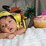 baby, yellow_hat, cupcake, pink_frosting, sprinkles, blanket, white_blanket, face, infant, indoor, decorations, balloon, sun_illustration, curious, resting, soft_texture, celebration, birthday, closeup, child