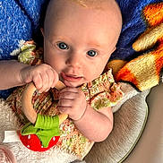 Winter is registered to the contest to win money with this photo: baby, infant, toy, wooden_ring, blanket, blue_blanket, orange_blanket, clothing, floral_dress, hand, face, eyes, skin, person, cute, portrait, indoors, soft_texture, child, teething_toy