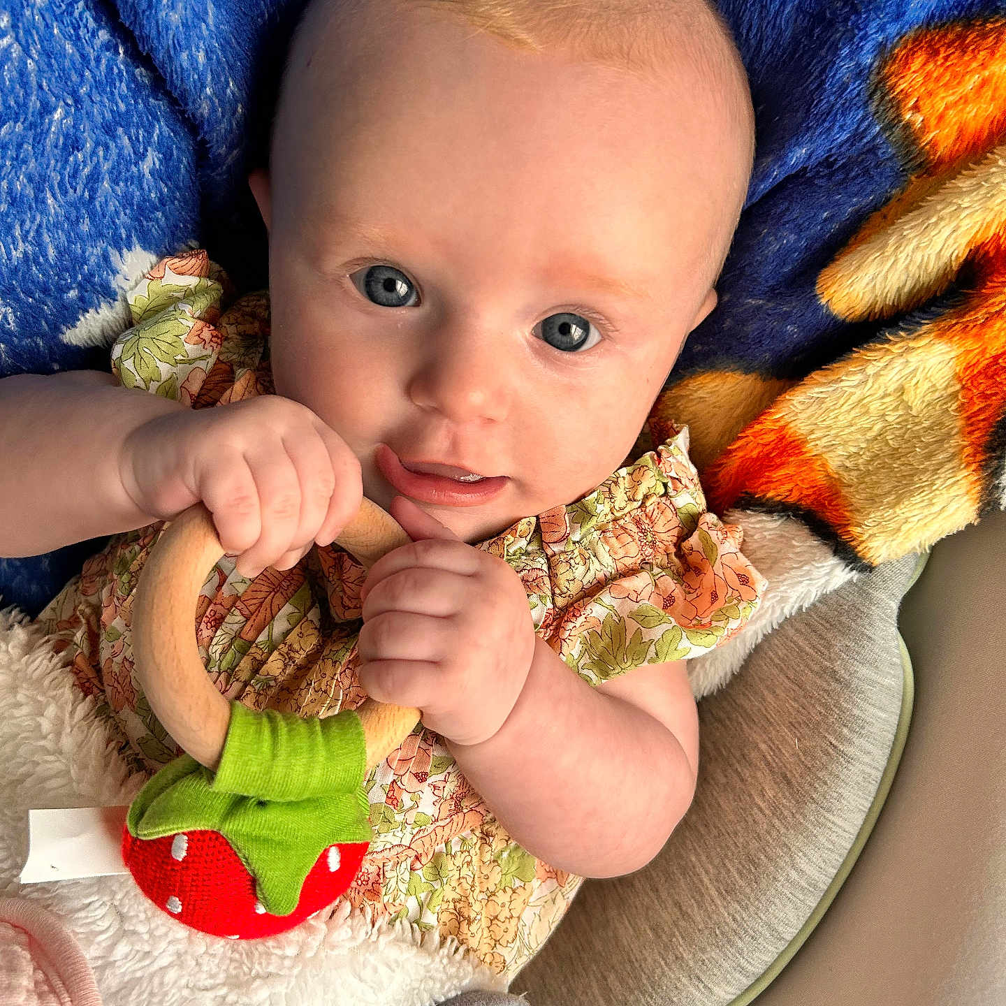 Winter is registered to the contest to win money with this photo: baby, infant, toy, wooden_ring, blanket, blue_blanket, orange_blanket, clothing, floral_dress, hand, face, eyes, skin, person, cute, portrait, indoors, soft_texture, child, teething_toy