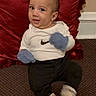baby, child, sitting, carpet, red_pillow, white_shirt, nike_logo, blue_mittens, black_pants, socks, cartoon_face, indoor, cute, young_child, floor, wall, expression, portrait, small_child, infant