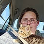 animal, car_interior, casual_clothing, cat, closeup, daytime, glasses, human_face, kitten, person, pet, safety, seatbelt, short_hair, sitting, sunlight, tabby_cat, transportation, travel, window