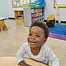 toddler, child, smiling, happy, table, classroom, education, toys, chair, floor, wall, cubicles, bag, curly_hair, long_sleeve, playroom, indoor, young_child, person, casual_clothing
