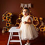 Melissa a rejoint le concours — aidez-le/la à gagner de superbes lots ! toddler, child, dress, white_shoes, stool, sunflowers, birthday, celebration, decorations, lights, bee_theme, brown_background, flooring, portrait, indoor, cute, standing, party, one_year, happy