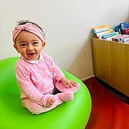 Beya participe au concours pour gagner de l'argent avec cette photo : baby, child, pink_clothing, headband, green_chair, smiling, indoor, bookshelf, books, floor, happy, person, seated, cute, infant, portrait, furniture, head, room, toy
