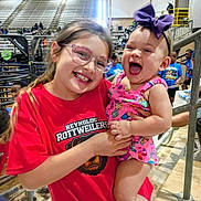 Kyndall is registered to the contest to win money with this photo: child, baby, girl, glasses, smiling, happy, sports_event, bleachers, crowd, red_shirt, purple_bow, holding, people, indoor, casual_clothing, person, cheerful, young_child, playful, family