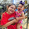 child, baby, girl, glasses, smiling, happy, sports_event, bleachers, crowd, red_shirt, purple_bow, holding, people, indoor, casual_clothing, person, cheerful, young_child, playful, family