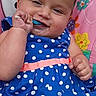 Nova is registered to the contest to win money with this photo: baby, child, blue_dress, polka_dot, spoon, smiling, sitting, colorful_background, floral_pattern, infant, cute, happy, face, hand, seat, indoors, portrait, food, feeding, young_child