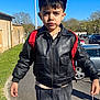 Göktuğ a rejoint le concours — aidez-le/la à gagner de superbes lots ! child, boy, outdoor, leather_jacket, black_cap, backpack, pavement, trees, cars, sunny, daytime, street, sidewalk, young, person, standing, holding, casual_clothing, sky, urban