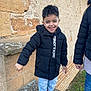 child, boy, smiling, jacket, black_jacket, jeans, sneakers, outdoor, stone_wall, standing, happy, casual_clothing, daylight, person, fashion, young, playful, fun, portrait, urban