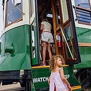 Murphy is registered to the contest to win money with this photo: child, toddler, curly_hair, dress, tram, vintage, green, adult, shorts, steps, outdoor, transportation, vehicle, side_view, daylight, person, walking, street, public_transport, curious
