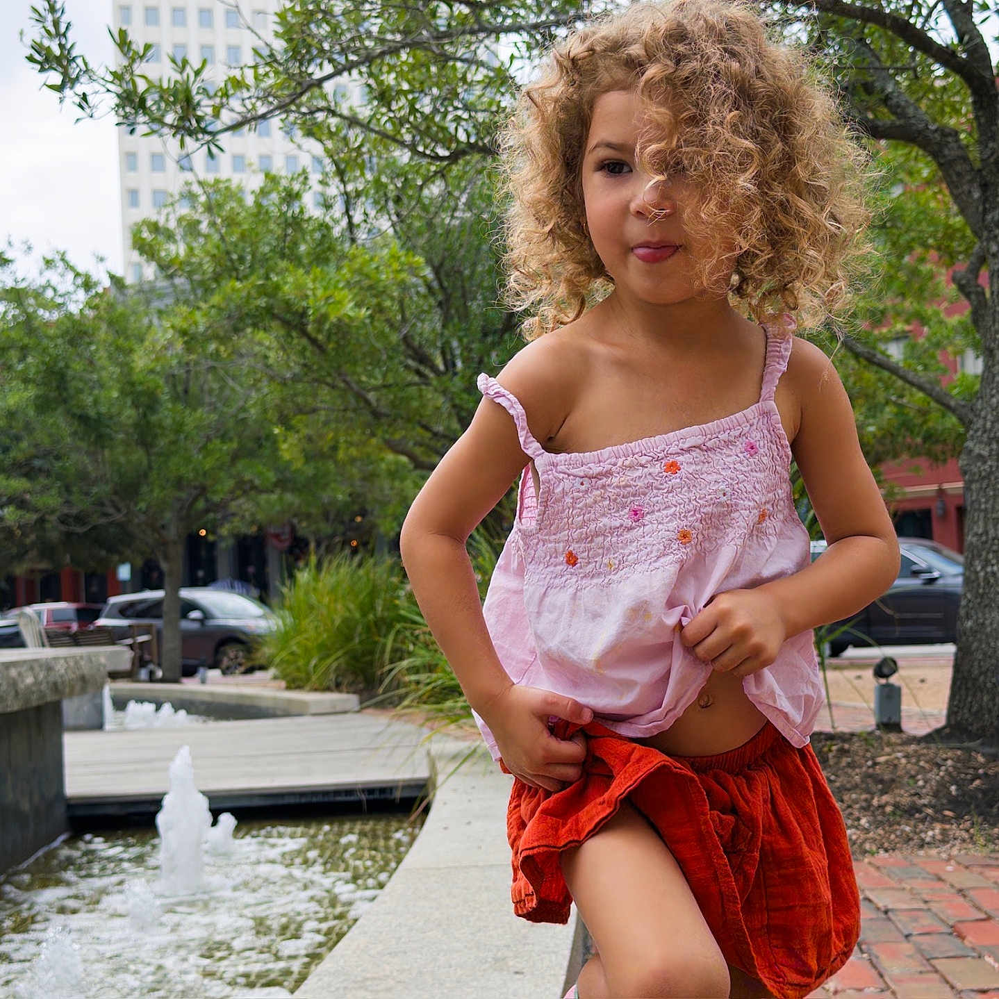 Murphy joined the competition — help win amazing prizes! building, child, curly_hair, daylight, fountain, greenery, happy, nature, orange_shorts, outdoor, park, person, pink_top, playful, sidewalk, smiling, tongue_out, trees, urban, water