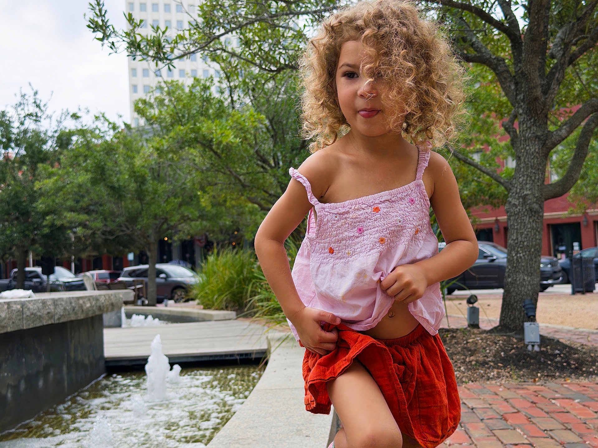 Murphy joined the competition — help win amazing prizes! child, curly_hair, pink_top, orange_shorts, fountain, water, trees, urban, sidewalk, playful, tongue_out, daylight, building, park, nature, person, outdoor, greenery, smiling, happy