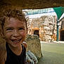 Murphy is registered to the contest to win money with this photo: child, smile, curly_hair, outdoor, playground, rock, stone, shade, joyful, happy, person, young, girl, earrings, black_dress, daylight, background, architecture, fun, candid