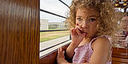 Murphy joined the competition — help win amazing prizes! child, toddler, curly_hair, pink_dress, thumb_sucking, window, wooden_panel, vehicle, reflection, person, indoor, seat, casual_clothing, portrait, face, expression, curiosity, daylight, candid, human