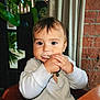Fox is registered to the contest to win money with this photo: toddler, child, baby, indoor, eating, snack, face, hands, curly_hair, long_sleeve_shirt, table, chair, plant, brick_wall, curtains, portrait, cute, seated, food, closeup