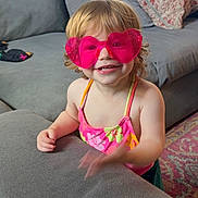 Elia is registered to the contest to win money with this photo: toddler, child, sunglasses, heart_shaped, pink, smiling, swimsuit, floral_pattern, couch, living_room, carpet, pillow, curtains, indoors, happy, cute, blond_hair, small_child, playful, furniture