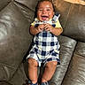 Zyrae is registered to the contest to win money with this photo: baby, blue_sandals, checkered_clothing, child, comfortable, cute, face, feet, hands, happy, indoors, infant, leather_couch, legs, person, relaxed, romper, sitting, smiling, young_child