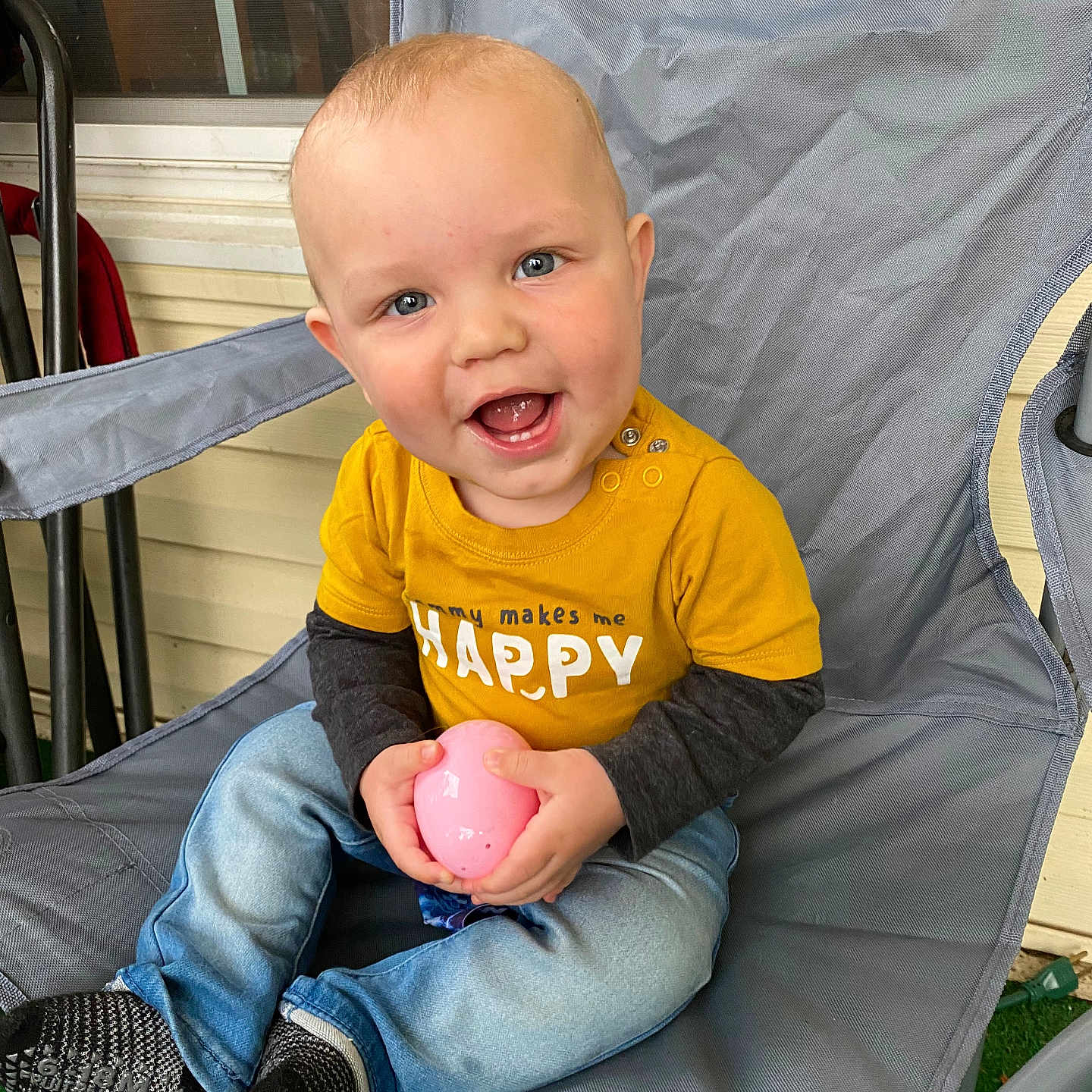 Jameson joined the competition — help win amazing prizes! baby, blue_eyes, casual_clothing, child, cute, expression, gray_chair, happy, holding_object, infant, jeans, outdoor, person, pink_egg, porch, seated, smiling, socks, yellow_shirt, young_child