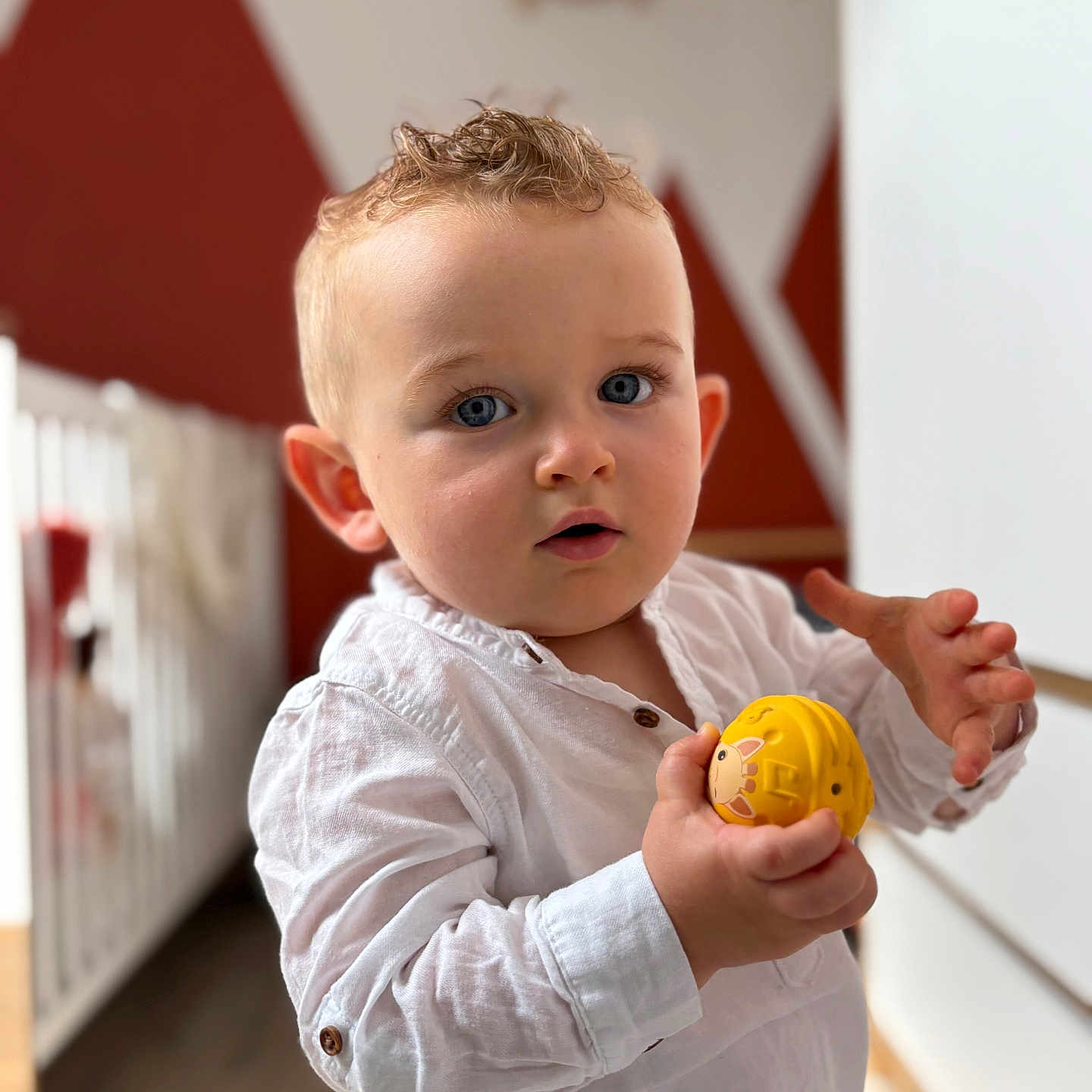 Ange participe au concours pour gagner de l'argent avec cette photo : blue_eyes, blurred_background, casual_clothing, child, crib, curly_hair, expression, face, hand, indoor, light, person, portrait, standing, toddler, toy, wall_art, white_shirt, wood_floor, yellow_toy