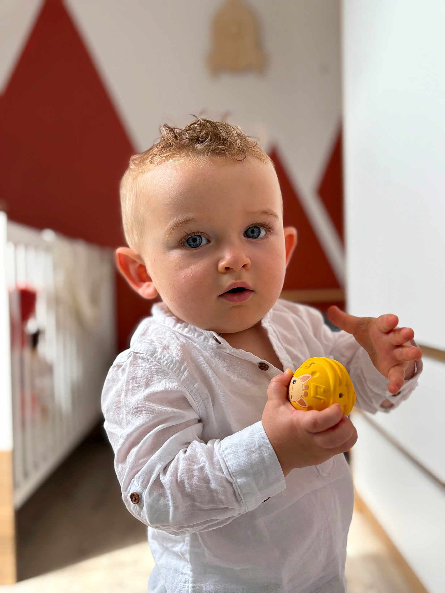 Ange participe au concours pour gagner de l'argent avec cette photo : blue_eyes, blurred_background, casual_clothing, child, crib, curly_hair, expression, face, hand, indoor, light, person, portrait, standing, toddler, toy, wall_art, white_shirt, wood_floor, yellow_toy