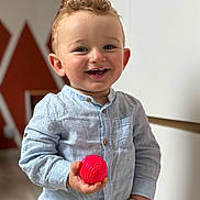 Ange participe au concours pour gagner de l'argent avec cette photo : ball, blue_shirt, buttoned_shirt, casual_clothing, child, curly_hair, floor, geometric_pattern, happy, holding_object, indoor, light_skin, modern_interior, person, playful, portrait, smiling, toddler, toy, young_child